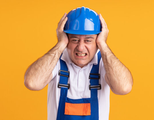 表情穿制服的未出租的成年建筑工人把手放在隔离在橙色墙上的头上姿势手制服