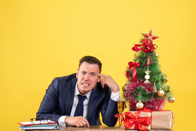 商务前视图愤怒的年轻人坐在圣诞树旁的桌子上 黄色背景上的礼物免费空间办公室职业新郎