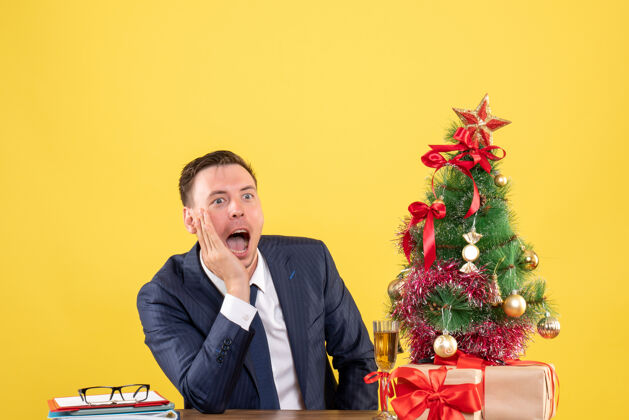生意前视图令人惊讶的男子大叫 而坐在圣诞树附近的桌子上 黄色背景上的礼物西装坐着人