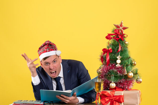 工作前视图愤怒的商人戴着圣诞帽坐在圣诞树旁的桌子旁 黄色背景上的礼物生意人礼物人