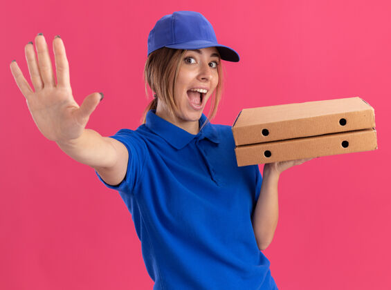感觉快乐的年轻漂亮的女送货员穿着制服 手里拿着比萨饼盒 伸出一只孤立在粉红色墙上的手伸展人制服