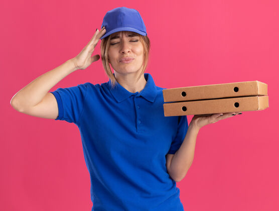 表情穿着制服的年轻漂亮的女送货员疼痛地把手放在头上 手里拿着隔离在粉红色墙上的比萨饼盒人漂亮感觉