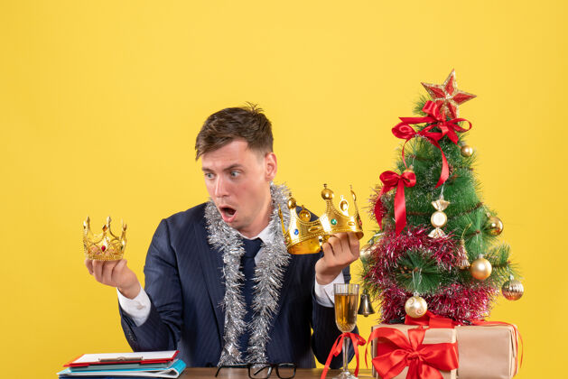 人前视图惊奇的商人看皇冠坐在圣诞树附近的桌子和黄色背景上的礼物树前面人