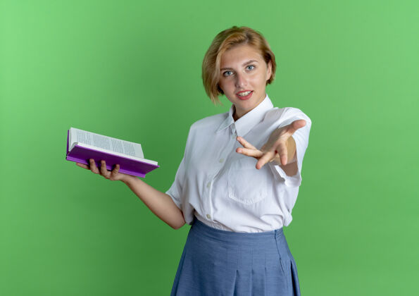 自信年轻自信的金发俄罗斯女孩拿着书 向隔离在绿色背景上的相机伸出手来 并留有复印空间金发手女孩
