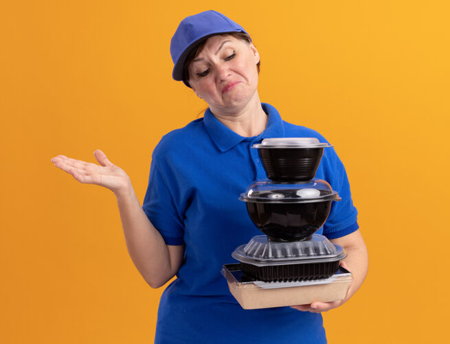 困惑身穿蓝色制服 戴着帽子的中年送货妇女拿着一叠食品包装 困惑地看着他们 不高兴地举起手臂站在橙色的墙上女人食物帽子