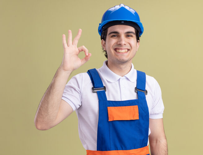 年轻人年轻的建筑工人穿着建筑制服 戴着安全帽 微笑着看着前面 绿色的墙上挂着“ok”的牌子制服安全微笑