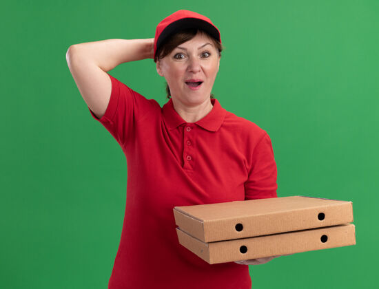 制服中年送货员身穿红色制服 头戴鸭舌帽 拿着披萨盒 站在绿色的墙边惊讶地看着前面盒子拿着惊喜