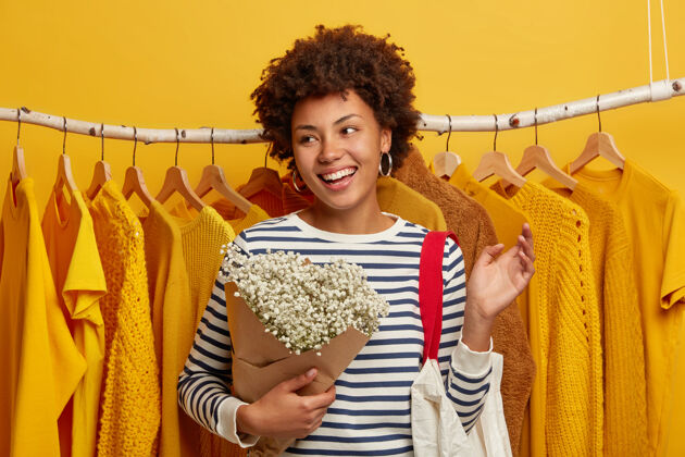 购买乐观的女顾客在商店里购物 拿着花束和包在架子上的黄色衣服上摆姿势 微笑着聚精会神地站在一边时尚请衣柜