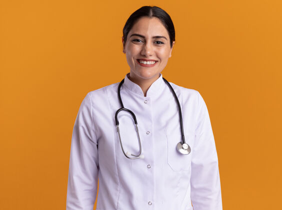 快乐快乐的年轻女医生 穿着医用外套 脖子上戴着听诊器 站在橘色的墙上 面带微笑地看着前方站大外套