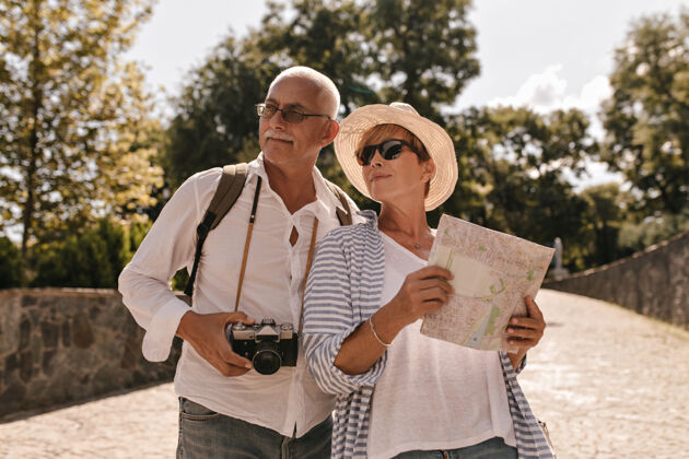 情感戴着帽子的短发女士 戴着很酷的太阳镜 穿着蓝色条纹衣服 拿着地图 在公园里和戴眼镜 穿着白衬衫 带着相机的男人合影祖父母聪明老人