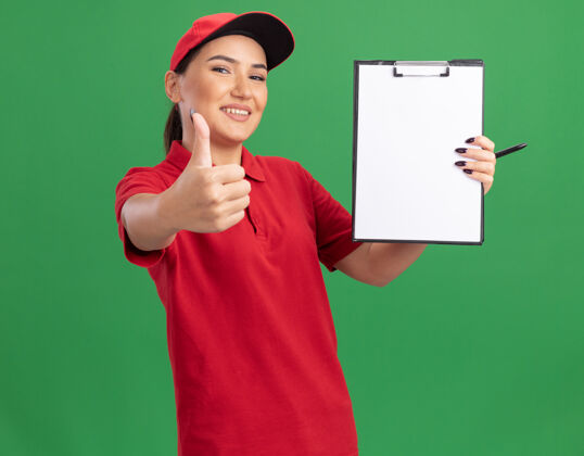 年轻身穿红色制服 头戴帽子的年轻送货员拿着空白页的剪贴板 面带微笑地站在绿色的墙上竖起大拇指页面女人帽子