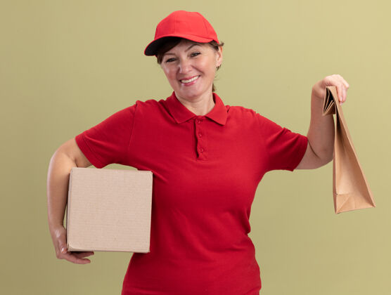 盒子快乐的中年女送货员穿着红色制服 戴着帽子 拿着纸包和纸板箱 站在绿色的墙边 微笑着看着前面包装给予纸板