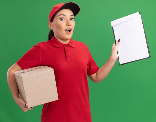 页面年轻的送货员身穿红色制服 头戴鸭舌帽 手里拿着纸板箱和写字板 手里拿着空白的书页 惊讶地站在绿色的墙上看着前面纸板女人送货