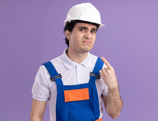 年轻年轻的建筑工人穿着建筑制服 戴着安全帽 看着前面 悲伤的表情困惑地指着站在紫色墙上的自己站着男人悲伤