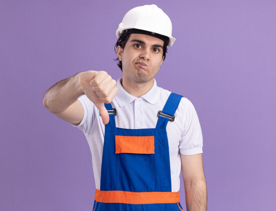 站起来年轻的建筑工人穿着建筑制服 戴着安全帽 不高兴地看着前面 竖起大拇指站在紫色的墙上大拇指不高兴制服