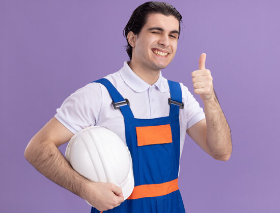 表情身穿建筑制服的年轻建筑工人举着安全帽 自信地微笑着看着前方 竖起大拇指站在紫色的墙上年轻建筑工人制服