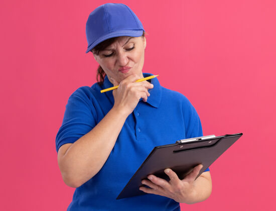 剪贴板身穿蓝色制服 头戴鸭舌帽 拿着剪贴板和铅笔的中年女送货员站在粉红色的墙上 神情迷茫 非常焦虑帽子焦虑困惑