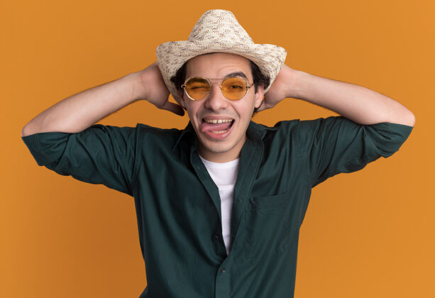 疯狂疯狂快乐的年轻人穿着绿色衬衫 戴着夏天的帽子 戴着眼镜 看着前面伸出的舌头站在橙色的墙上舌头年轻夏天