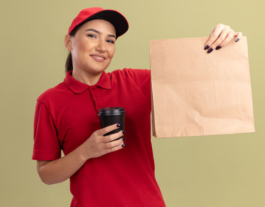 快乐身着红色制服 头戴鸭舌帽 拿着纸包和咖啡杯 脸上挂着微笑 站在绿色墙壁上的快乐年轻女送货员帽子女人脸