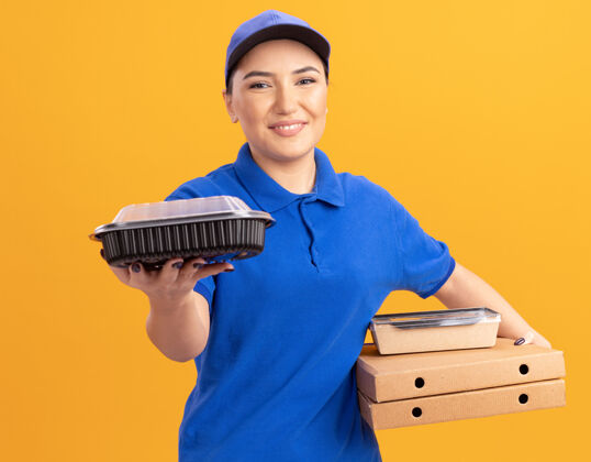 制服快乐的年轻送货员身穿蓝色制服 头戴鸭舌帽 拿着披萨盒和食品包 微笑着自信地站在橙色的墙上看着前方送货拿着年轻