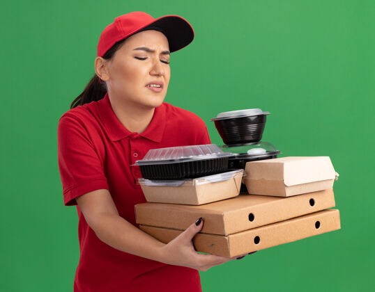 盒子年轻的送货员身穿红色制服 头戴鸭舌帽 手里拿着披萨盒和食品包 站在绿色的墙上 看上去既困惑又不高兴困惑送货制服