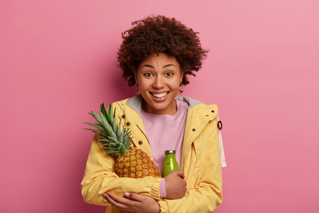 粉彩快乐的卷发女人的肖像拥抱新鲜多汁的菠萝和绿色水果冰沙 穿着黄色的休闲服 积极地微笑高兴姿势成分