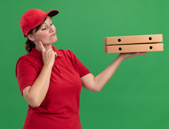 站着身穿红色制服 头戴鸭舌帽的中年送货妇女站在绿色的墙上 面带沉思的表情看着比萨饼盒表情女人中年