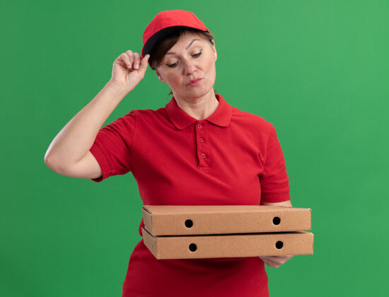 制服身穿红色制服 头戴鸭舌帽的中年送货妇女站在绿色的墙上 严肃地看着披萨盒披萨脸盒子