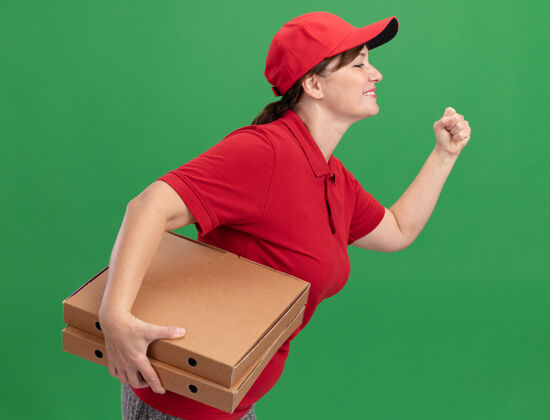 顾客身着红色制服 头戴鸭舌帽的快乐女送货员正在绿色墙壁上为顾客运送比萨饼盒跑步送货盒子