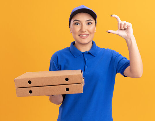 送货身穿蓝色制服 头戴鸭舌帽 拿着披萨盒 做着小动作的年轻女送货员站在橙色的墙上 面带微笑地看着前面做拿着帽子