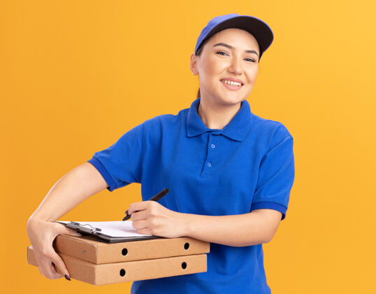 剪贴板身穿蓝色制服 戴着便签簿和拿着铅笔的披萨盒的年轻送货员站在橙色的墙上 面带微笑地看着前面拿着微笑欢呼