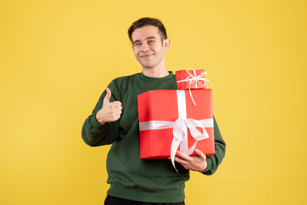 黄色正面图：年轻人拿着圣诞礼物 竖起大拇指 站在黄色背景上向上肖像休闲