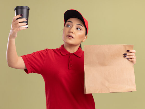 制服身穿红色制服 头戴鸭舌帽的年轻女送货员拿着纸包 另一只手端详着咖啡杯 严肃的脸站在绿色的墙上包装递送站着