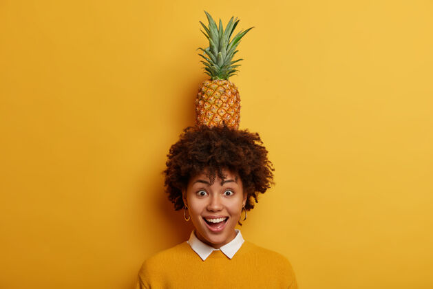 少女看我能做什么快乐搞笑的非洲发型女人头上抱着多汁的菠萝 开心大笑头发美国牙齿