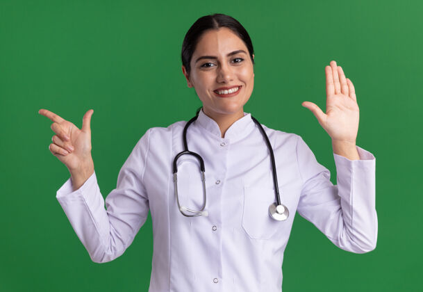 目录身穿医用外套的年轻女医生 脖子上戴着听诊器 看着前面抬起的手臂 食指指向一边 微笑着站在绿色的墙上指着外套壁板