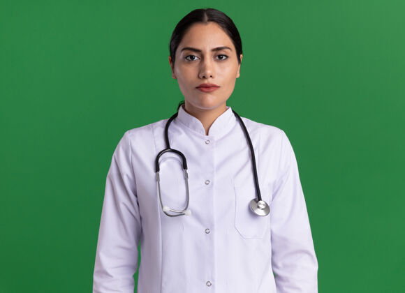 女人身穿医用外套 脖子上戴着听诊器的年轻女医生站在绿墙上 神情严肃自信地看着前方周围站严肃
