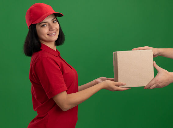 年轻身穿红色制服 头戴鸭舌帽的年轻送货员站在绿色的墙上 微笑着向顾客赠送纸板箱帽子给予站立