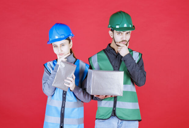 工人戴着头盔的男女工程师手持银色礼盒 神情迷茫 若有所思激动年轻人