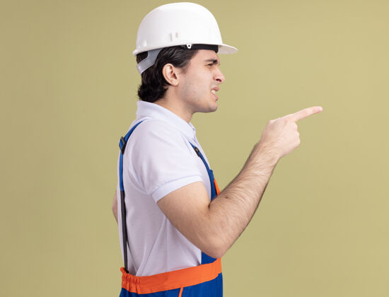 头盔年轻的建筑工人穿着建筑制服 戴着安全帽 用食指指着被迷惑的一边 站在绿色的墙上手指人建设者
