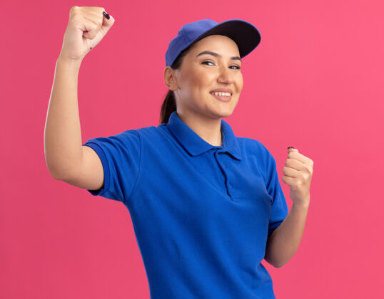 女人身穿蓝色制服 握紧帽子的年轻女送货员站在粉红色的墙上 快乐而兴奋制服交货握紧