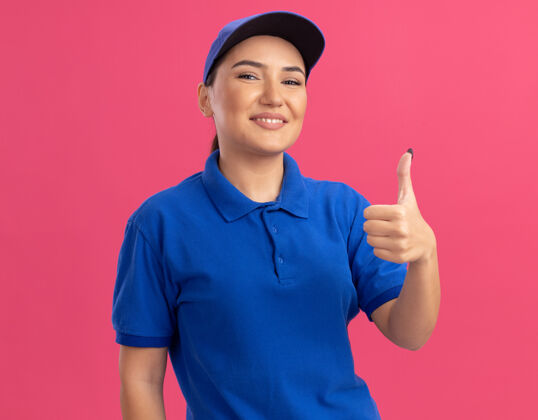 交货身穿蓝色制服 头戴鸭舌帽的年轻女送货员站在粉红色的墙上 自信地微笑着竖起大拇指看着前面立场女人信心