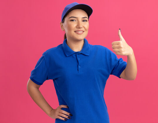 拇指身穿蓝色制服 头戴鸭舌帽的年轻女送货员站在粉红色的墙上 自信地微笑着竖起大拇指看着前面帽子微笑显示