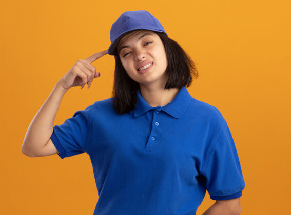 头穿着蓝色制服 戴着帽子的年轻送货女孩正困惑地用手指在橘色的墙上搔头站立制服帽子