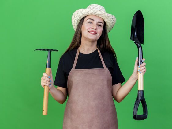 花园微笑美丽的园丁女孩穿着制服戴着园艺帽子拿着耙子和铁锹隔离在绿色背景上女孩园艺耙子