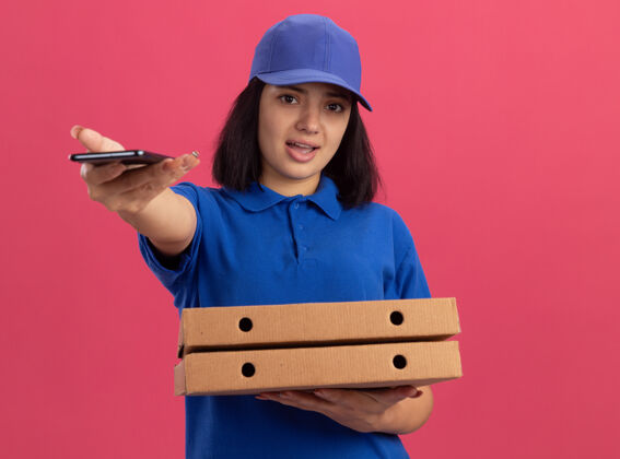拿着身穿蓝色制服 头戴鸭舌帽 手持披萨盒 伸出手机的年轻送货女孩站在粉红色的墙上伸展送货年轻人
