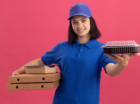 帽子身穿蓝色制服 头戴鸭舌帽 拿着比萨饼盒和食品包的年轻送货员微笑着站在粉红色的墙上食物拿着年轻人