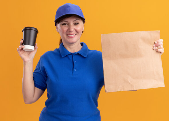 立场身着蓝色制服 戴着帽子的中年送货妇女拿着纸包 展示着咖啡杯 站在橙色的墙上 面带微笑地看着前面显示年龄交货