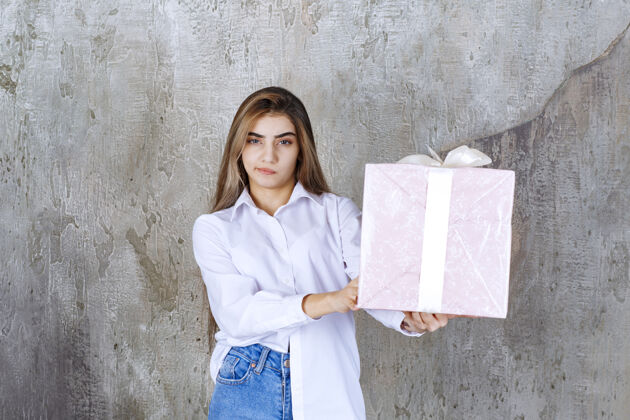 年轻人一个穿着白衬衫的女人手里拿着一个用白丝带包着的粉色礼盒 看上去困惑和犹豫思考庆祝礼盒
