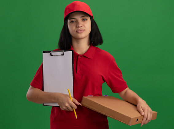 女孩身穿红色制服 头戴鸭舌帽 手持披萨盒和铅笔 脸上带着自信微笑的年轻送货女孩站在绿色的墙上递送拿着站着