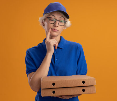女人身穿蓝色制服 戴着眼镜 拿着比萨饼盒的年轻送货员脸上挂着沉思的表情 望着橙色的墙上沉思帽子表情
